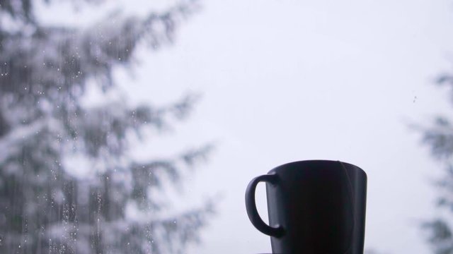 Teacup next to a winter when it is snowing outside.