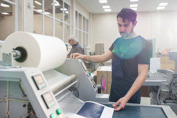 Serious young printing specialist with beard operating printing press while printing test page at...