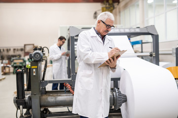 Serious senior inspector in lab coat standing at printing press with roll paper and making notes in...