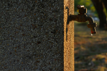 Old exterior water tap was closed and rough textured of polished stone wall with water leakage and sunrise or sunset lighting. Background for waterworks or drought problem concept. Selective focus.