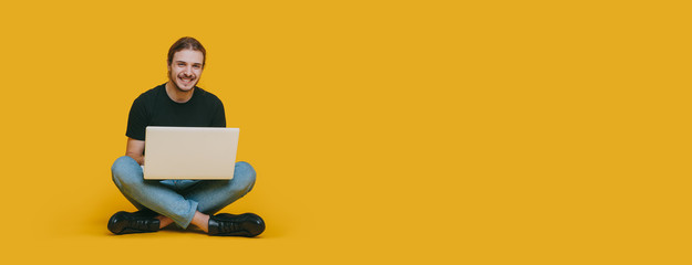 A happy boy with long hair and brown beard sitting with a computer on a yellow background with blank space