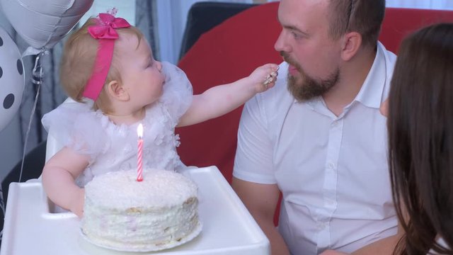 Family portrait mom, dad and baby girl at first birthday with cake and candle on it. Girl is eating cake by hands. Small home party with decorations. Happy smiling people. Family lovely moments.