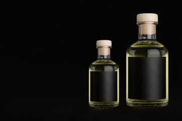 Big and small glass bottles for cosmetic, perfume, drink with black label, cork, yellow liquid on black background, mock up for design of product.