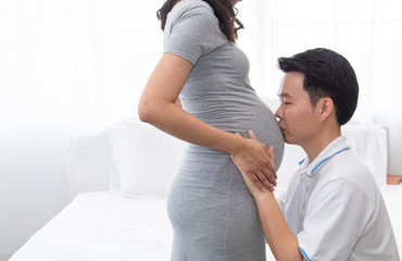 Beautiful Asian pregnancy woman, husband hold belly, tummy with love, kiss, caring togetherness on bed at home, excited motherhood awaiting for childbirth sharing moment with couple, family concept