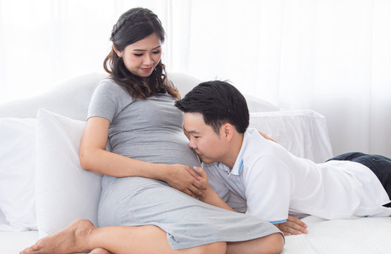 Beautiful Asian pregnancy woman, husband hold belly, tummy with love, kiss, caring togetherness on bed at home, excited motherhood awaiting for childbirth sharing moment with couple, family concept