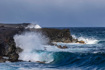 Wave crashing on black volcanic cliffs, black sand beach on Hawaii's Big Island. Ocean and blue sky in background. 