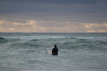 View of lonely woman surfer sitting on surfboard and looking towards sunset clouds on horizon
