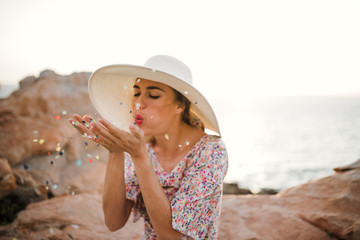 Beautiful woman with large summer hat blowing confetti on the beach