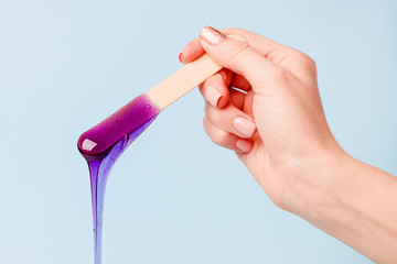 Hand holding beauty purple wax or sugar paste on wooden spatula flowing down into container on blue...