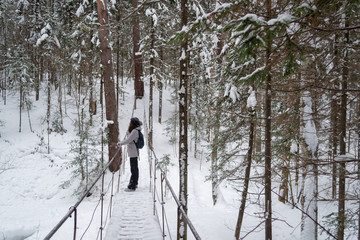 Young woman stands on a suspension bridge in the middle of a winter forest and looks into the distance