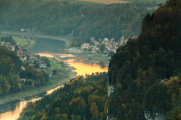 View of the village of Wehlen in the valley in the Saxon Switzerland National Park. River Elbe with trees and rocks in the autumn mood. Sunshine from the sunset is reflected in the water