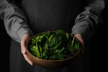 wooden plate with fresh spinach leaves in the hands of a woman dressed in gray linen clothes. closeup