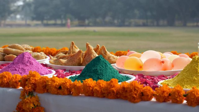Pan shot of a beautifully decorated festive table for Holi festival celebrated in India. Heaps of colorful Gulal with plates of samosas  Indian sweet Gujiyas  Kesar Thandai  and water bombs on the ...