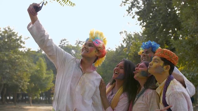 Young friends covered in Gulal colors taking selfies on a smartphone - Holi party. Group of excited Indian teenagers in traditional wear smiling and posing for pictures with colored faces during jo...