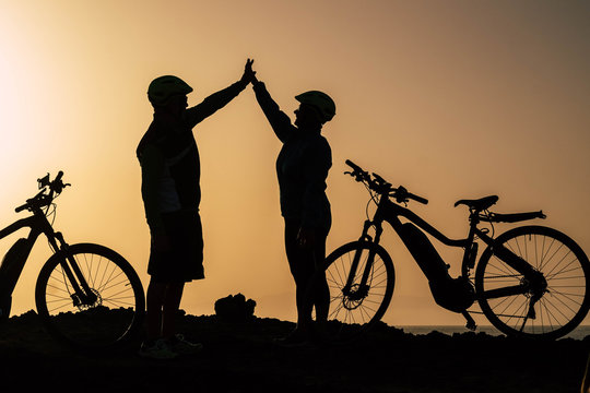 couple of two seniors and mature people together at the sunset with their bikes giving five with the silhouette and the sea at the background