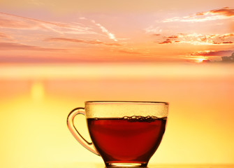 A transparent cup with black tea against the backdrop of a beautiful sunrise at sea. Water in the early morning sunlight.