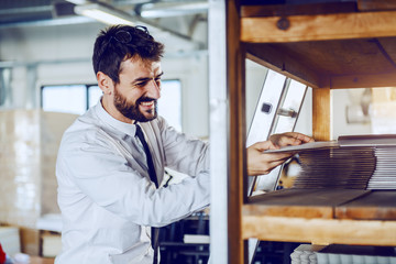 Fototapeta premium Smiling caucasian bearded supervisor in shirt and tie taking offset printing plate while standing in printing shop.