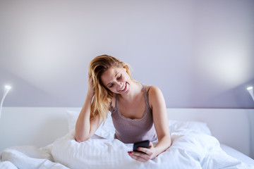 Attractive caucasian smiling blond woman sitting on bed in bedroom at night and reading message on smart phone.