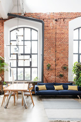 Loft apartment with big window in industrial style. Living room interior with design table with chairs and navy sofa with pillows. Vertical.