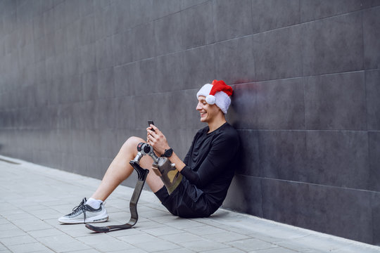 Cheerful Caucasian sportsman with artificial leg and with santa hat on head sitting on ground, leaning on wall and sending wishes for christmas over his smart phone.