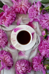 White cup with black tea or coffee with chocolate and petals on a saucer surrounded by fresh pink peonies, a wonderful morning or a coffee break. Flat lay, close up