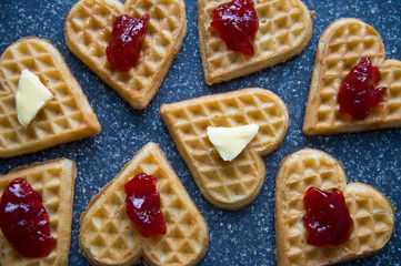 Group of heart-shaped waffles topped with berry jam and butter arranged on a grey stone background
