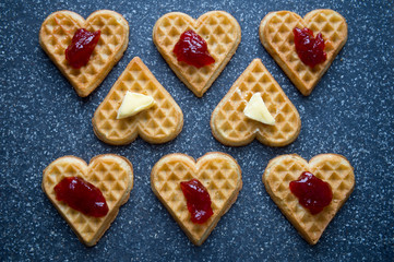 Group of heart-shaped waffles topped with berry jam and butter arranged on a grey stone background