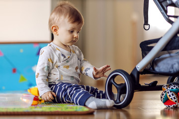 Adorable blond toddler sitting on the floor and playing with stroller wheel. Home interior.