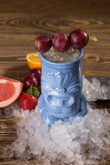 Tropical cocktail with grape in blue tiki style glass on crushed ice on wooden background