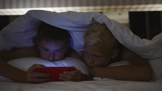 Children lie under a blanket at night and use the game app on their tablet.