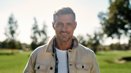 Beautiful sunny day. Portrait of handsome and stylish bearded man looking at camera and smiling while standing in the middle of the park