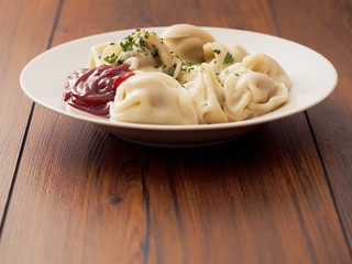 Portion of Russian classic big size dumplings on a white plate with tomato ketchup and herbs. Dark wood table surface.