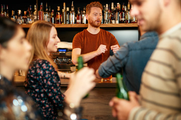 Cheerful friends leaning on bar counter, drinking beer and chatting with bartender. Night out.