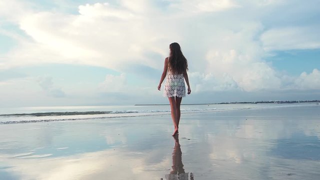 Woman walks barefoot along beach at sunset