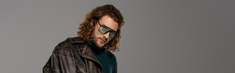 panoramic shot of handsome man in leather jacket looking at camera isolated on grey