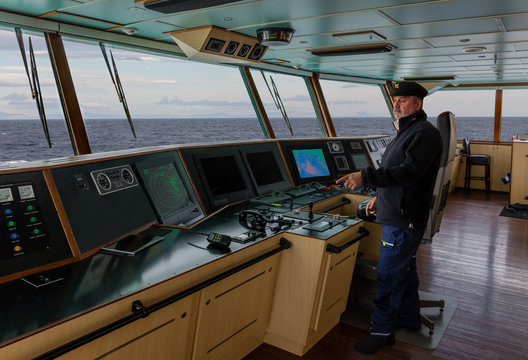 The captain with a beard and a blue cap stands on the bridge of a cargo ship and points to the handheld radio. Through the windows you can see the sea and mountains on the horizon.