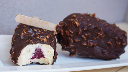 Two chocolate nuts and berry ice cream on a stick. One is whole, another is bitten and you can see berry filling. White plate. Grey background. 