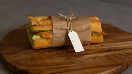 Salmon sandwich with avocado and cucumber wrapped in paper situated on wooden board. There is white card for inscription on a paper. Grey background.