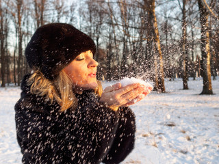 A young blonde woman in a fur coat and hat made of mink fur blows off light fluffy snow from the...