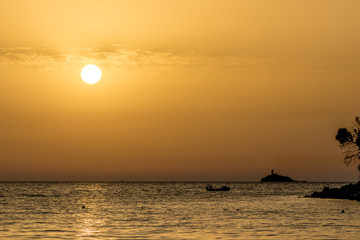 Focus on round setting Sun. Boat silhouette. Sun circle at beach at sunset in front of golden Ionian Sea water with slight overcast clouds. Dusk as seen from Ksamil, Albania, spring scenery evening