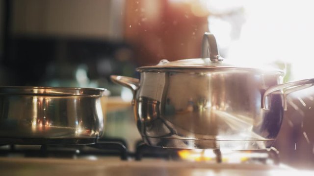 A cooking pot with a glass lid stands on a gas stove, boiling water in it. Steam and drops splash out. Solar glare from the kitchen window, soft focus