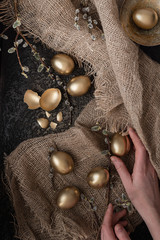 Golden Easter eggs are scattered on a dark background with burlap and willow twigs. female hands collect eggs.