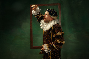 Shocked. Portrait of medieval young man in vintage clothing with wooden frame on dark background....
