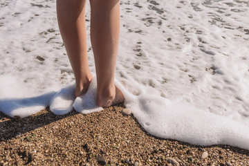 Closeup view of two small kid legs standing on sunny beach. Soft white foamy sea waves splashing around bare feet of child.