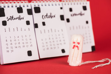Selective focus of hygienic tampon with sad face expression near calendar on red background