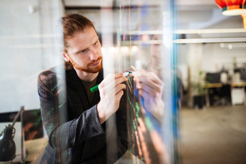 Young man working with datas and diagrams, writing ideas on glass office wall