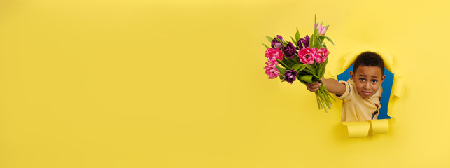 Smiling African-American with a bouquet of Tulip flowers looks out of torn paper gives flowers on yellow background. Concept of Valentine's Day, mother's Day, eighth of March, spring