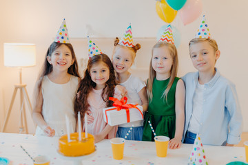 Glad birthday girl stands with presenet, happy friends come to congratulate her, wear party cone hats, stand near festive table with cake, smile joyfully, celebrate festive event