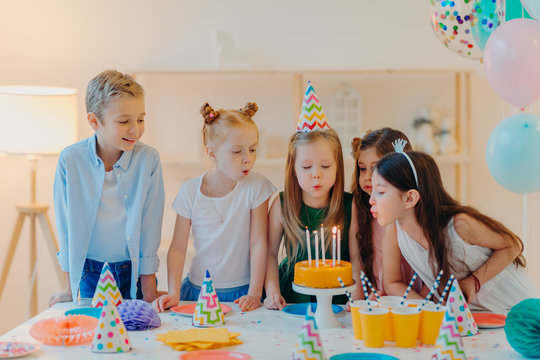 Small kids celebrate birthday party, blow candles on cake, gather at festive table, have good mood, enoy spending time together, make wish, wear party hats, pose indoor with inflated balloons