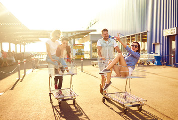 Young friends having fun on shopping trolleys together outdoor. Group of happy young people racing on shopping cart in summer day with sunlight. Lifestyle, leisure, entertainment, youth concept.